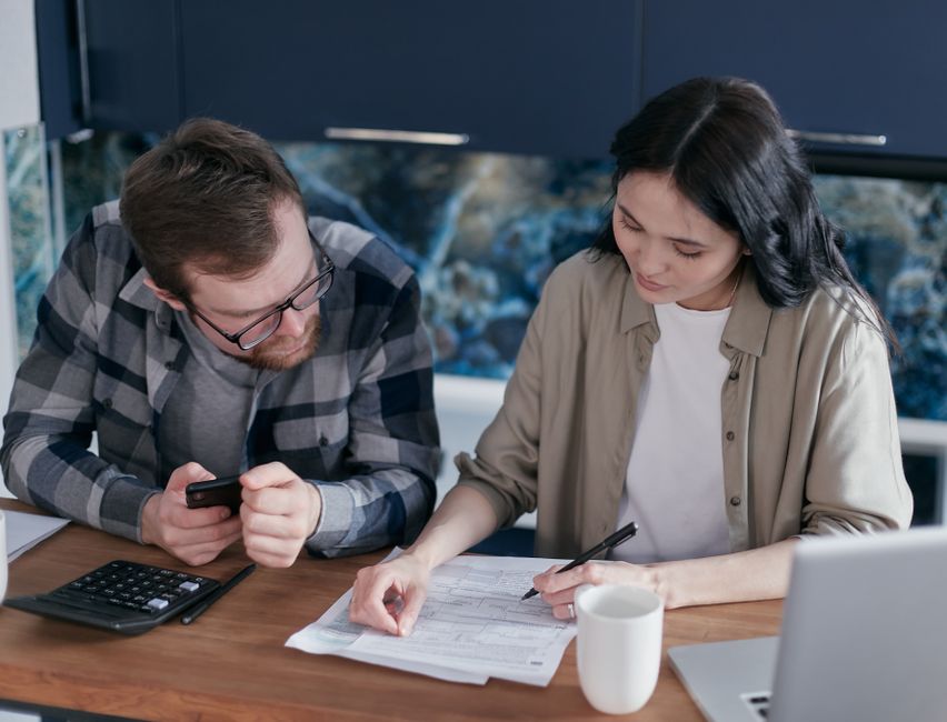 Professional couple sits at a wooden table in a bright, modern living room, carefully reviewing paperwork for their home p...
