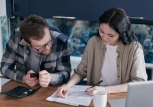 Professional couple sits at a wooden table in a bright, modern living room, carefully reviewing paperwork for their home p...